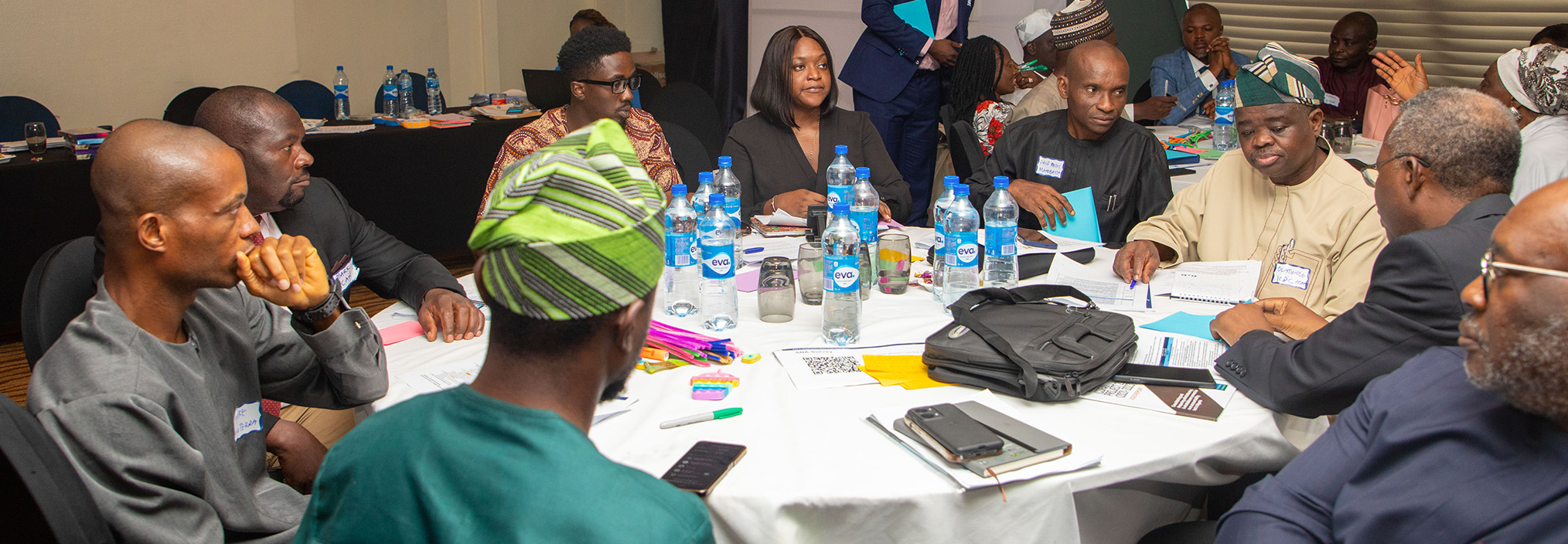 A group of people sit around a table in discussion at a meeting, with papers and water bottles on the table.